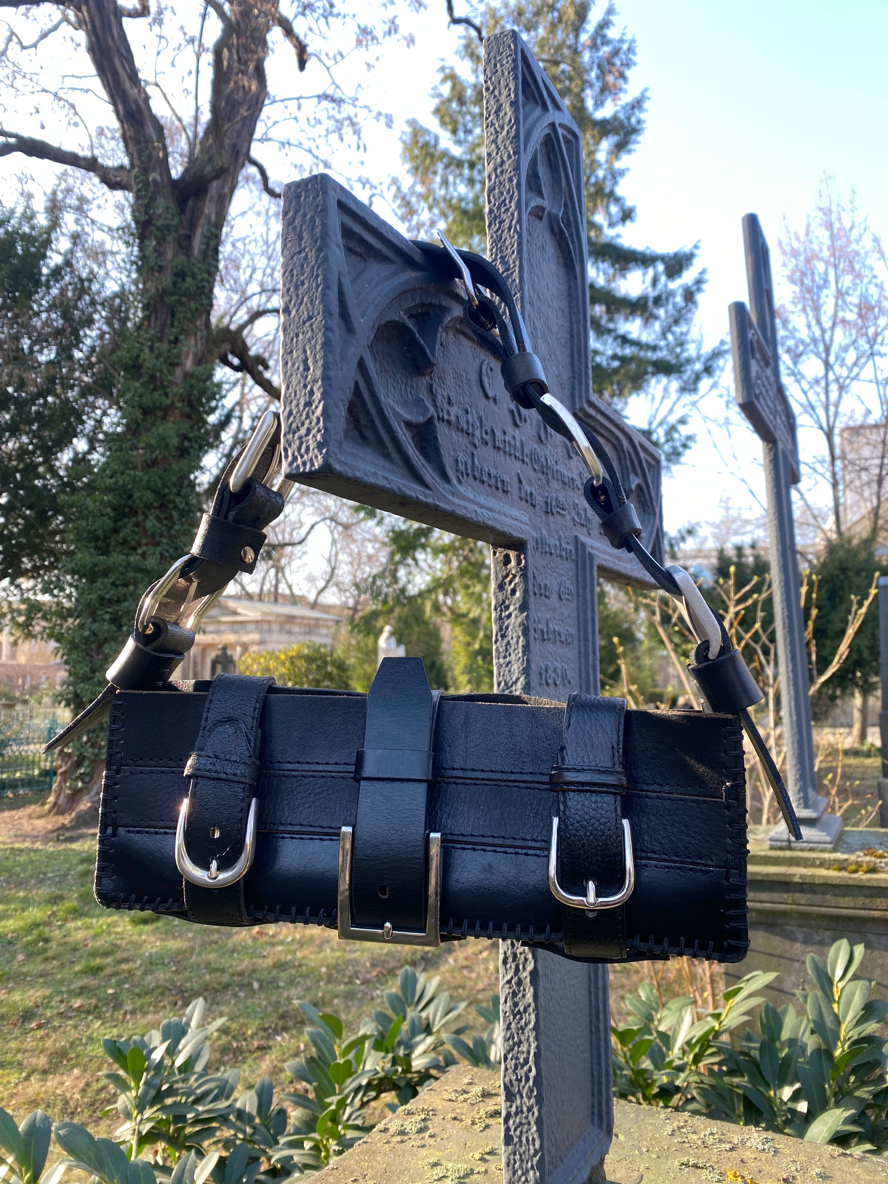 Long black leather-style handbag made from upcycled and repurposed materials, hanging on a metal cross for a gothic aesthetic. Eco-friendly, vegan, and sustainable shoulder bag design.