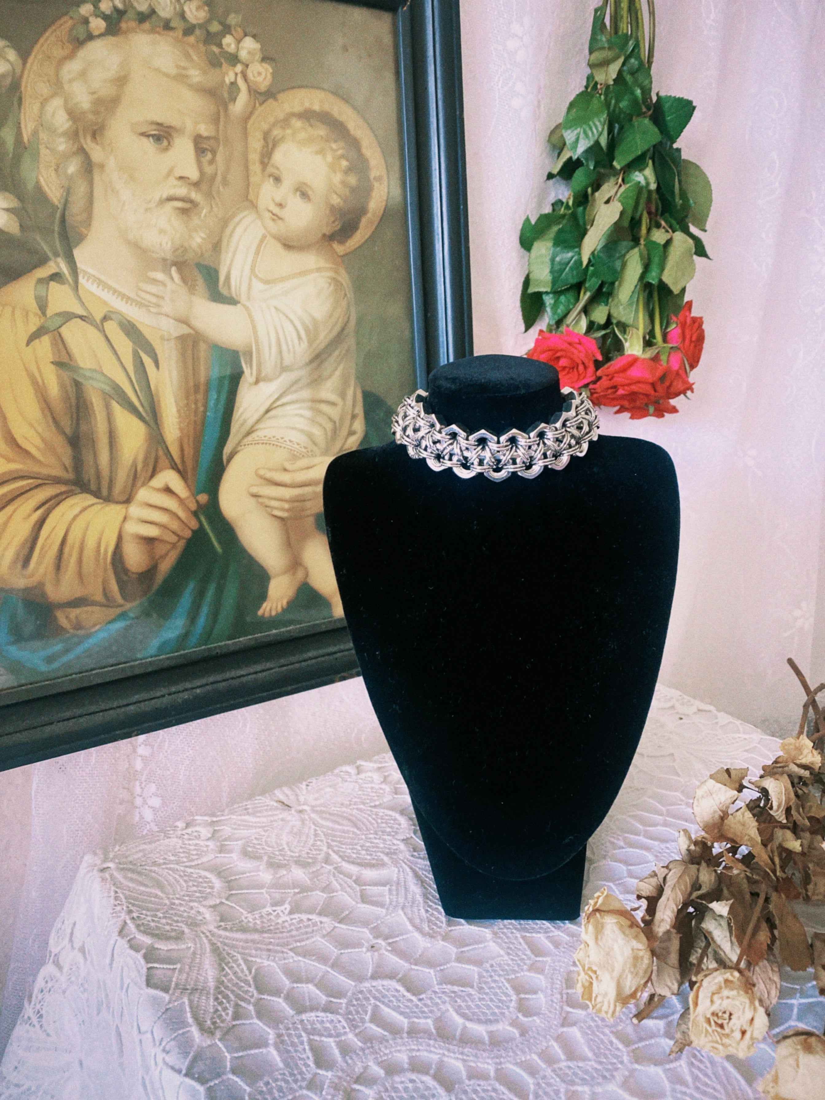 Industrial, heavy-duty handmade choker necklace made from stainless steel screws and chainmail, displayed on a doll with gothic aesthetics, accompanied by dried flowers and a Szent Kép, creating a gothic lookbook for alternative fashion.