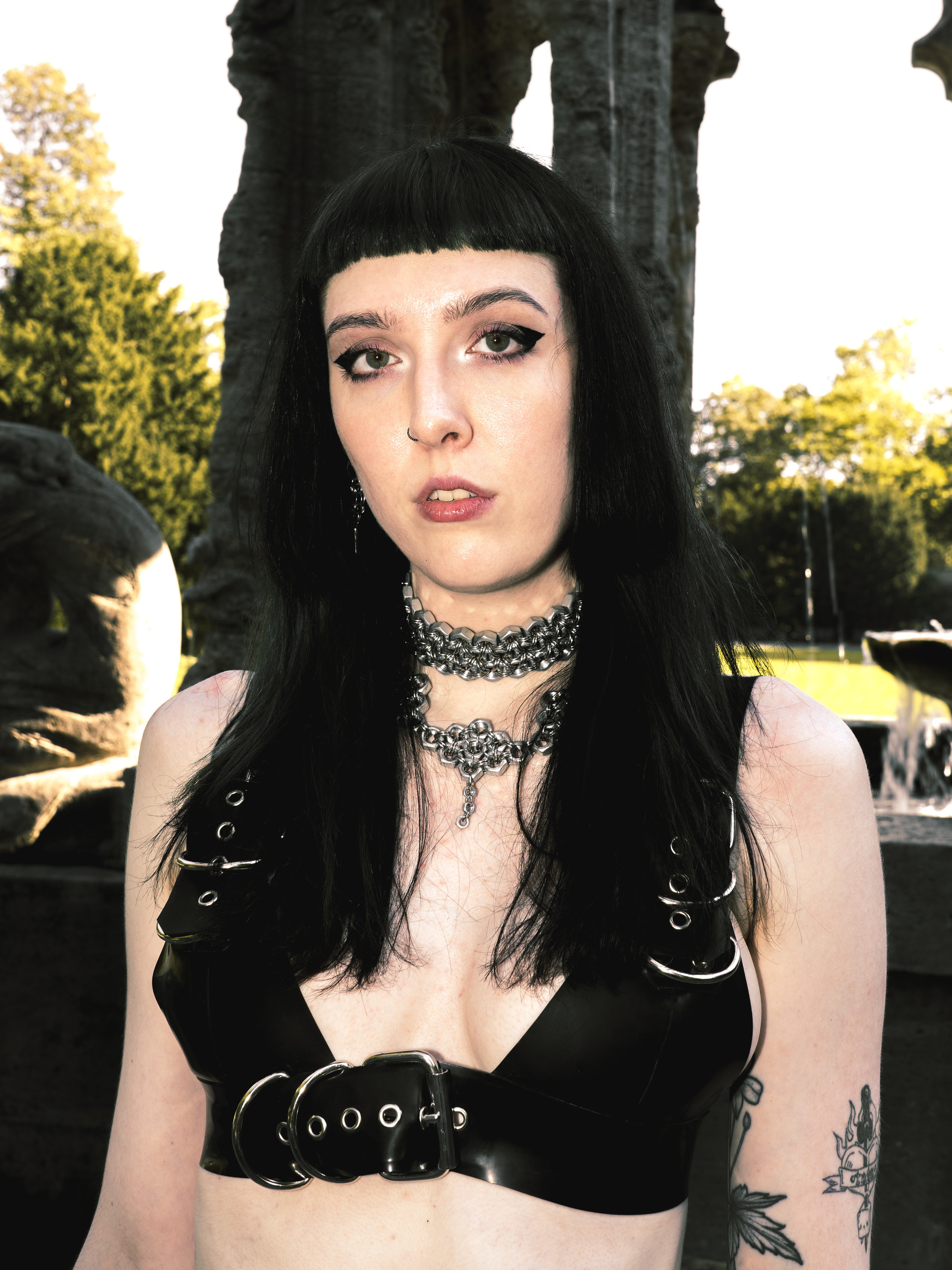 Model wearing a Triple-Row True Industrial Metal Choker Necklace made of screws and chainmail, styled with a latex bra in front of a gothic fountain, showcasing an alternative fashion aesthetic.