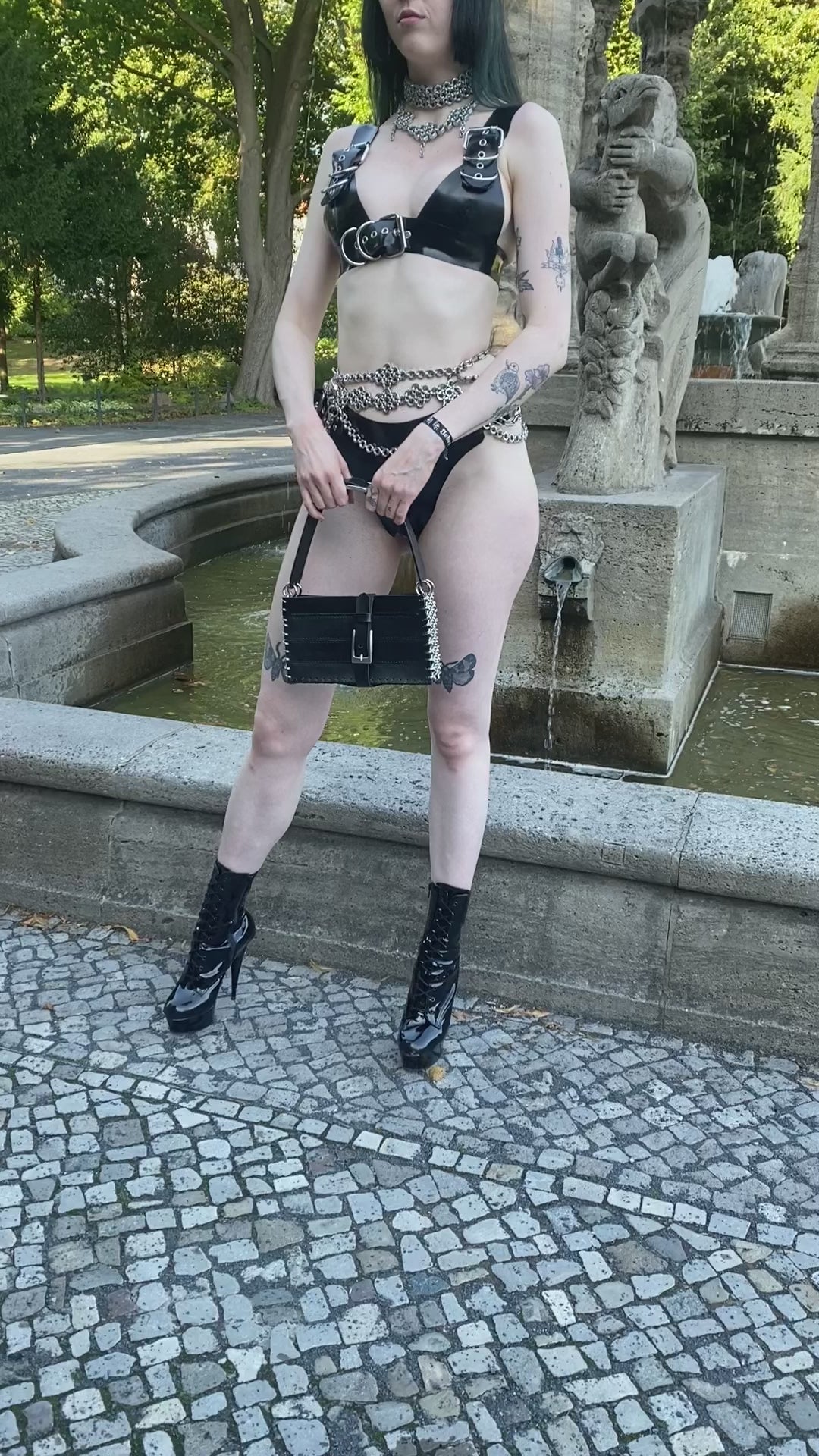 Alternative model wearing chainmail necklaces and black leather handbag with chainmail detail in black latex outfit in front of a gothic fountain for gothic aesthetics. Product made in Berlin.