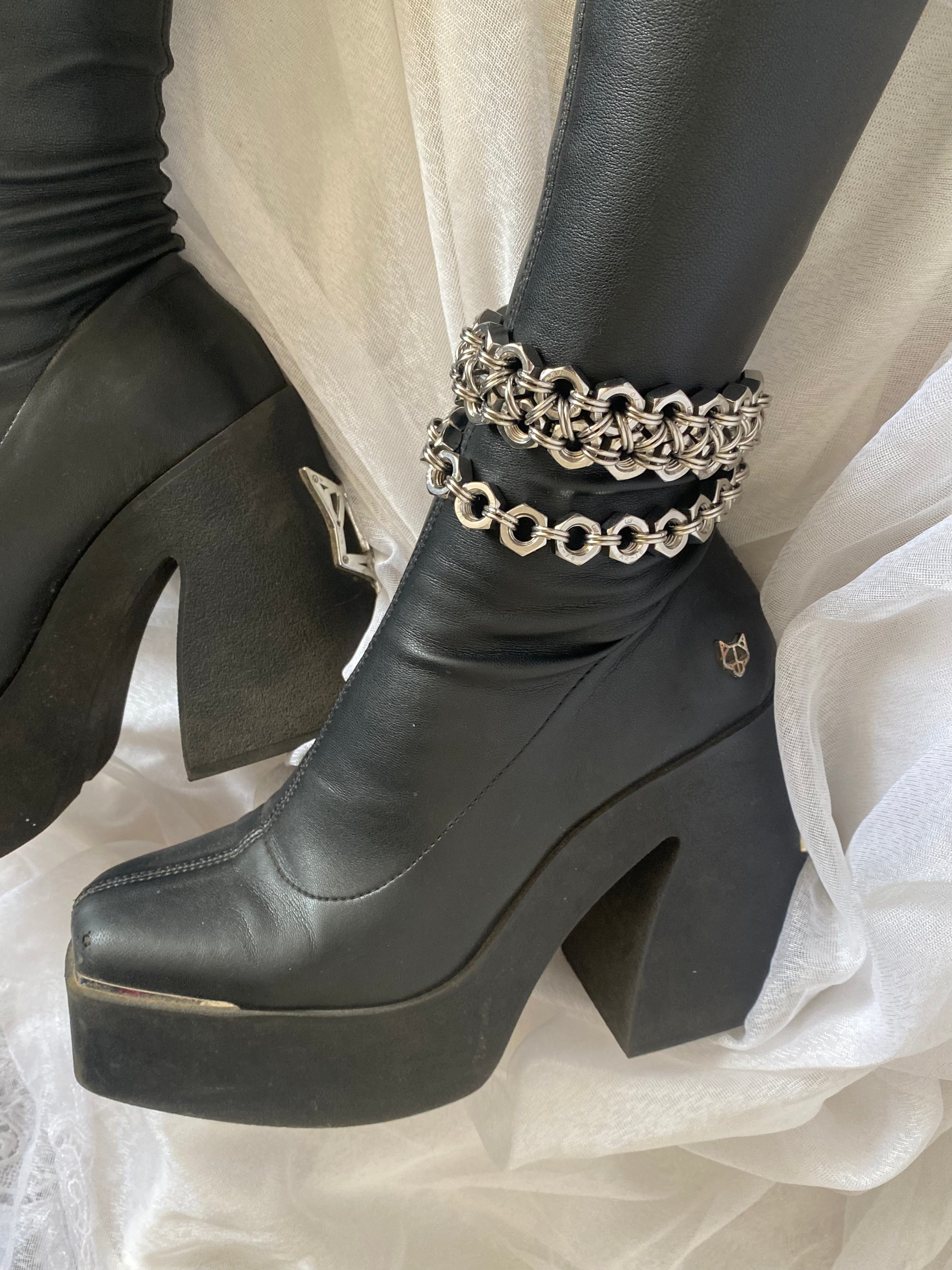 Industrial, slightly Celtic-style gothic chainmail boot charms, handcrafted from stainless steel, displayed on Naked Wolfe knee-high boots, in a gothic aesthetic, laying on white lace with tattooed legs.