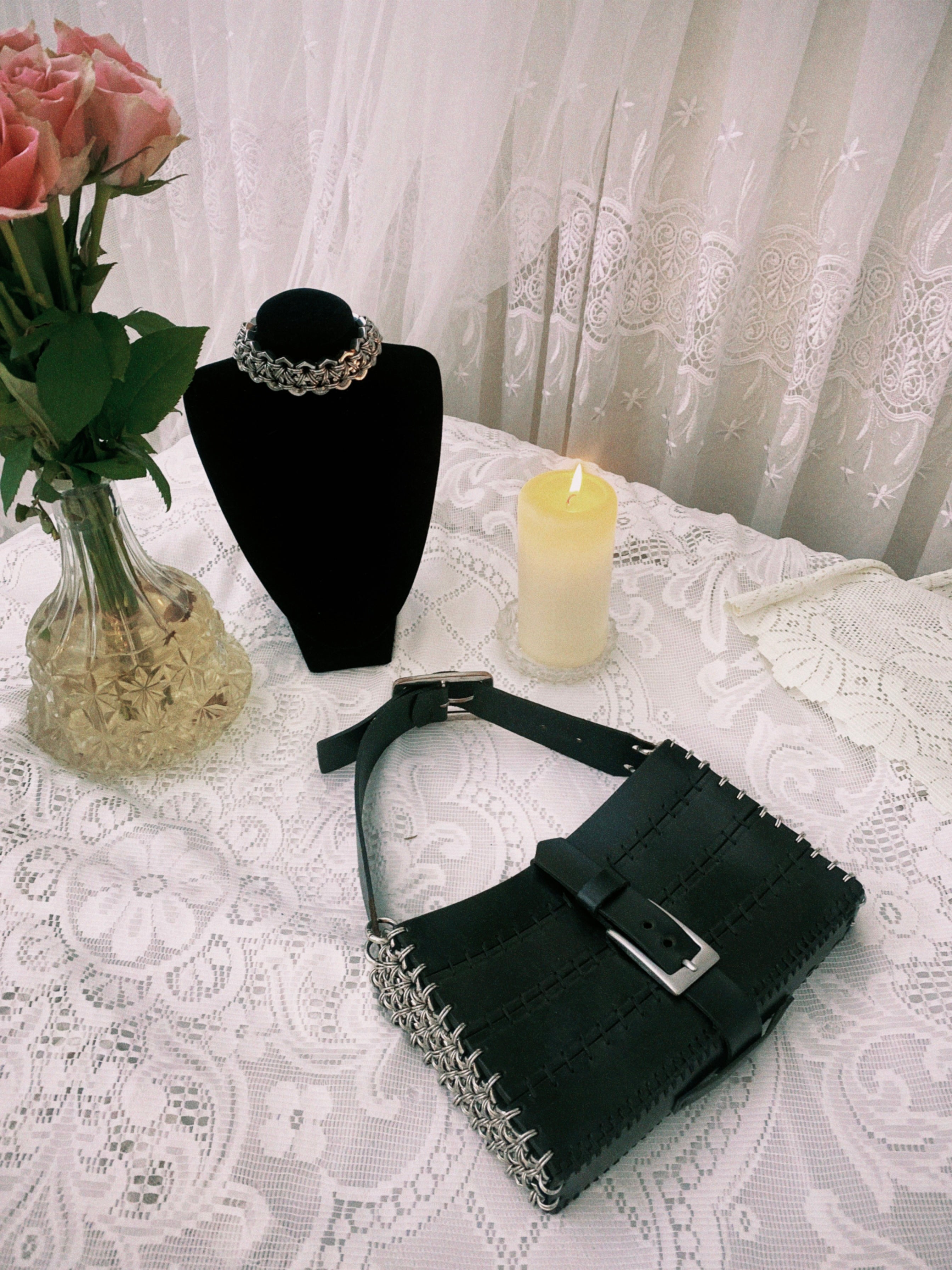 Heavy-duty stainless steel metal choker displayed with a black sustainable handbag featuring the same pattern; styled in a gothic setting with white lace, flowers, and candles.