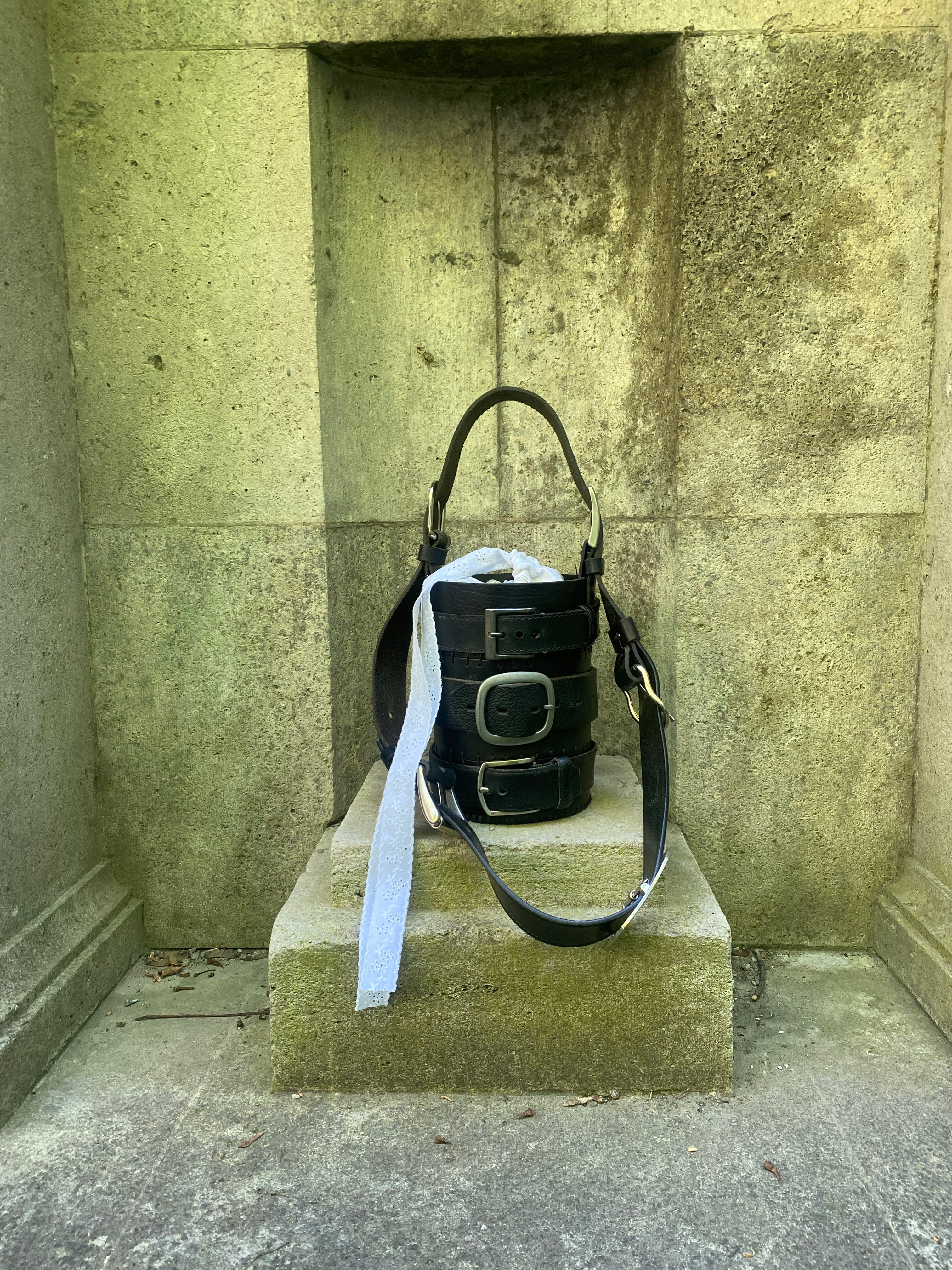 Cylinder-shaped black leather handbag made from repurposed leather and upcycled belt material, standing on a gothic-style rock surface. Bold statement design for fashion enthusiasts.