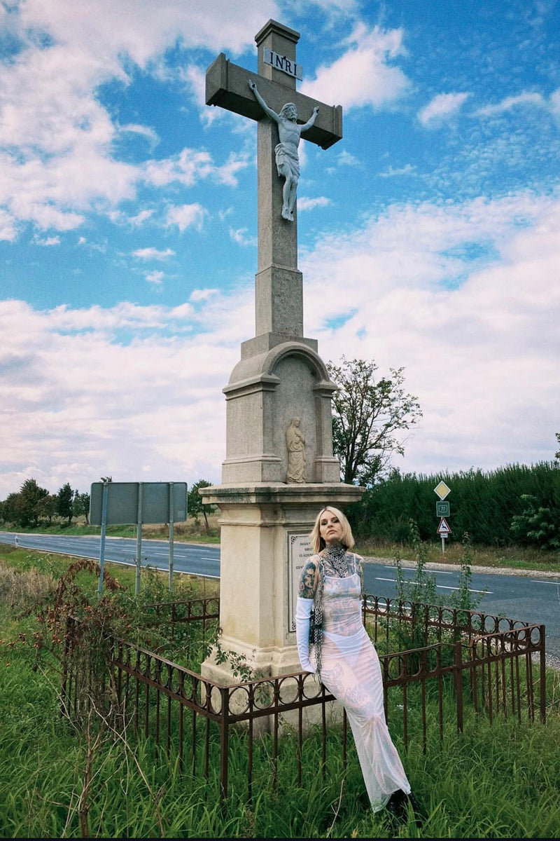 Tattooed woman in a sheer dress wearing a chainmail necklace and an upcycled leather handbag, sitting in front of a large cross at an intersection.