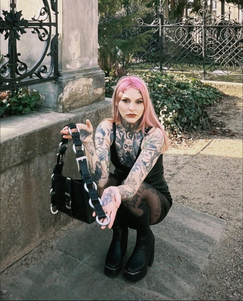Person with tattoos and pink hair holding a black repurposed leather handmade bag in a gothic setting.
