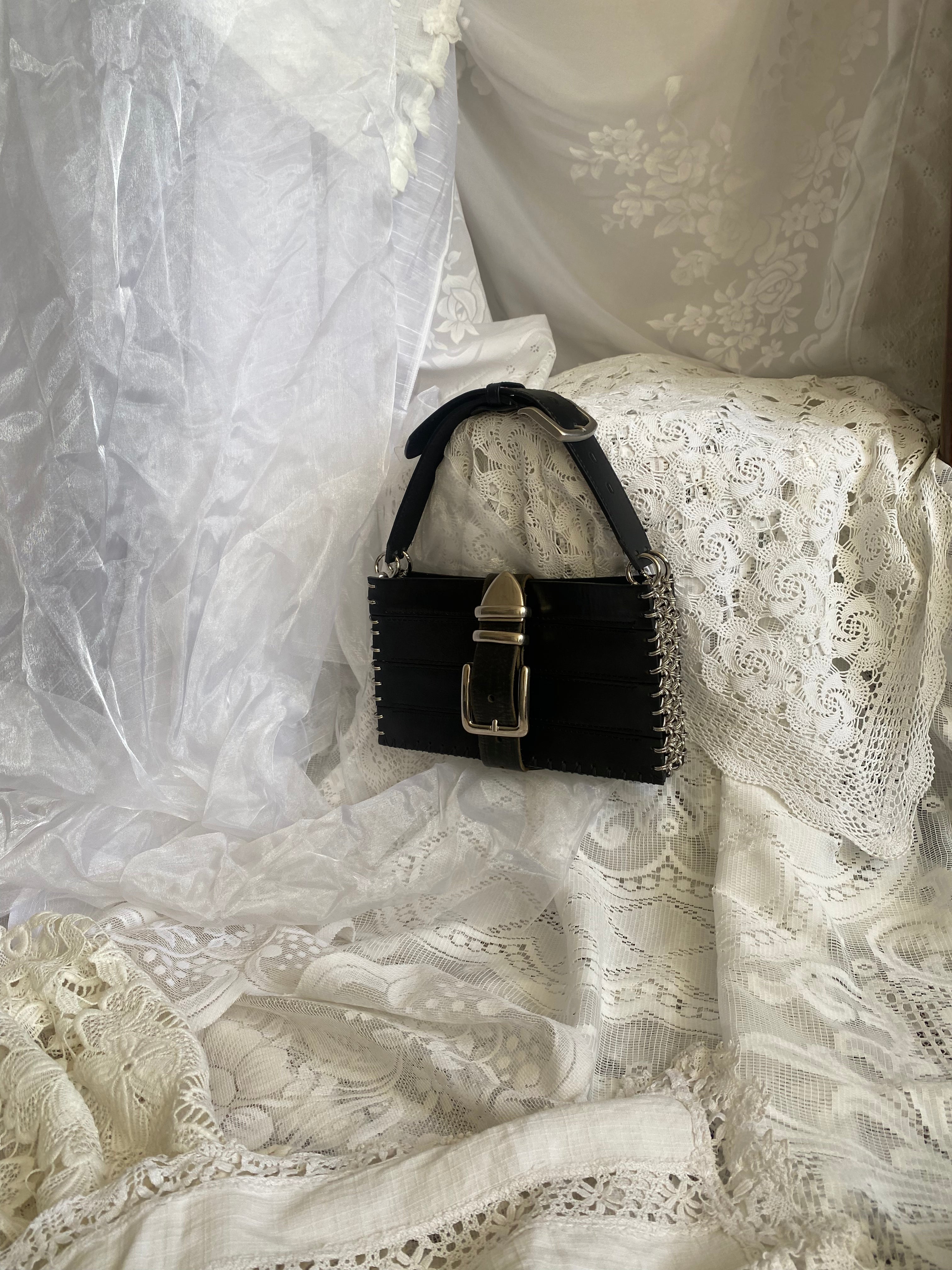 Black handbag crafted from upcycled leather, photographed against a gothic lace background. A statement piece shown from a distance, featuring machine-made panels and hand-stitched construction for exceptional durability.