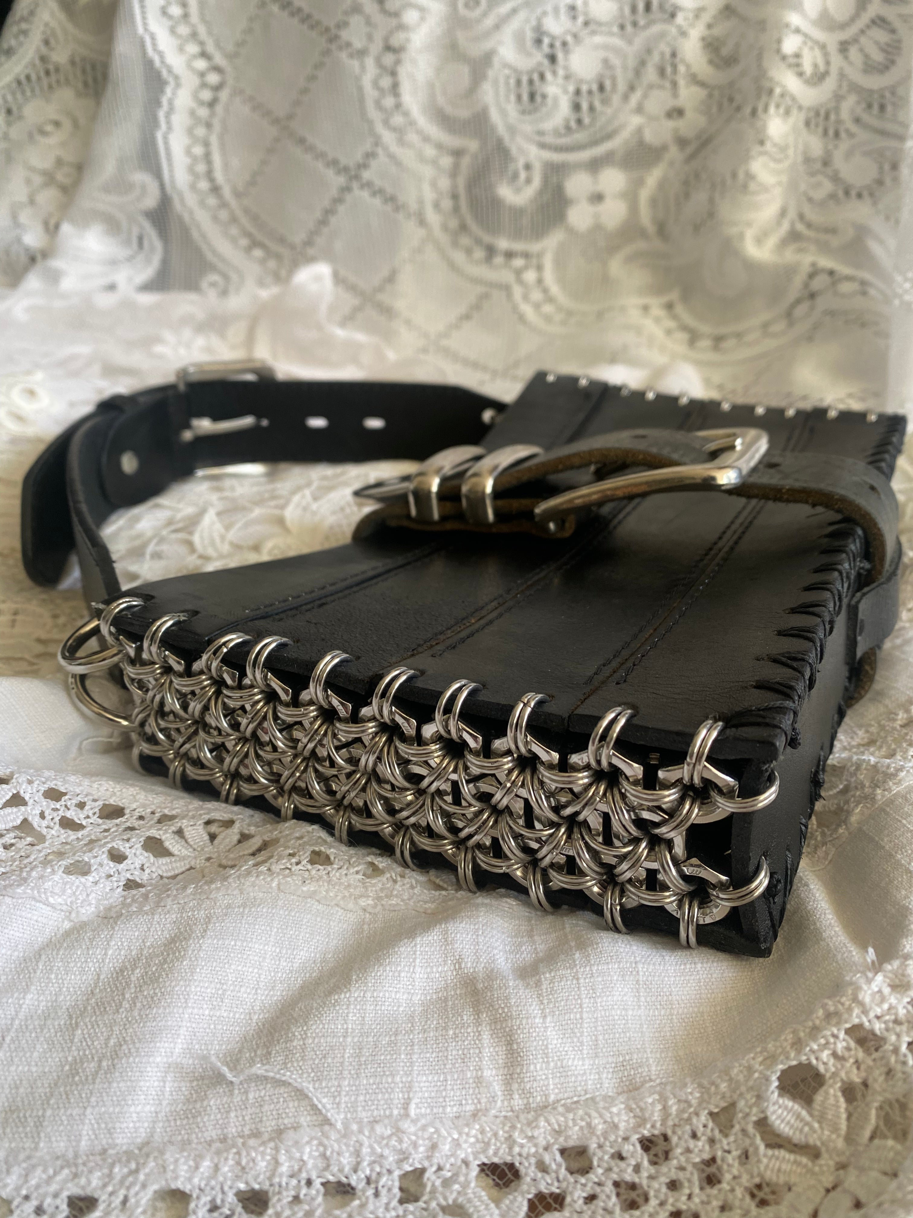 Limited edition handmade black leather bag, shown lying flat in a close-up that highlights bold chainmail side details made from stainless steel screws and chains. An avant-garde statement piece for bold wearers, photographed on white lace fabric for a gothic-inspired aesthetic.