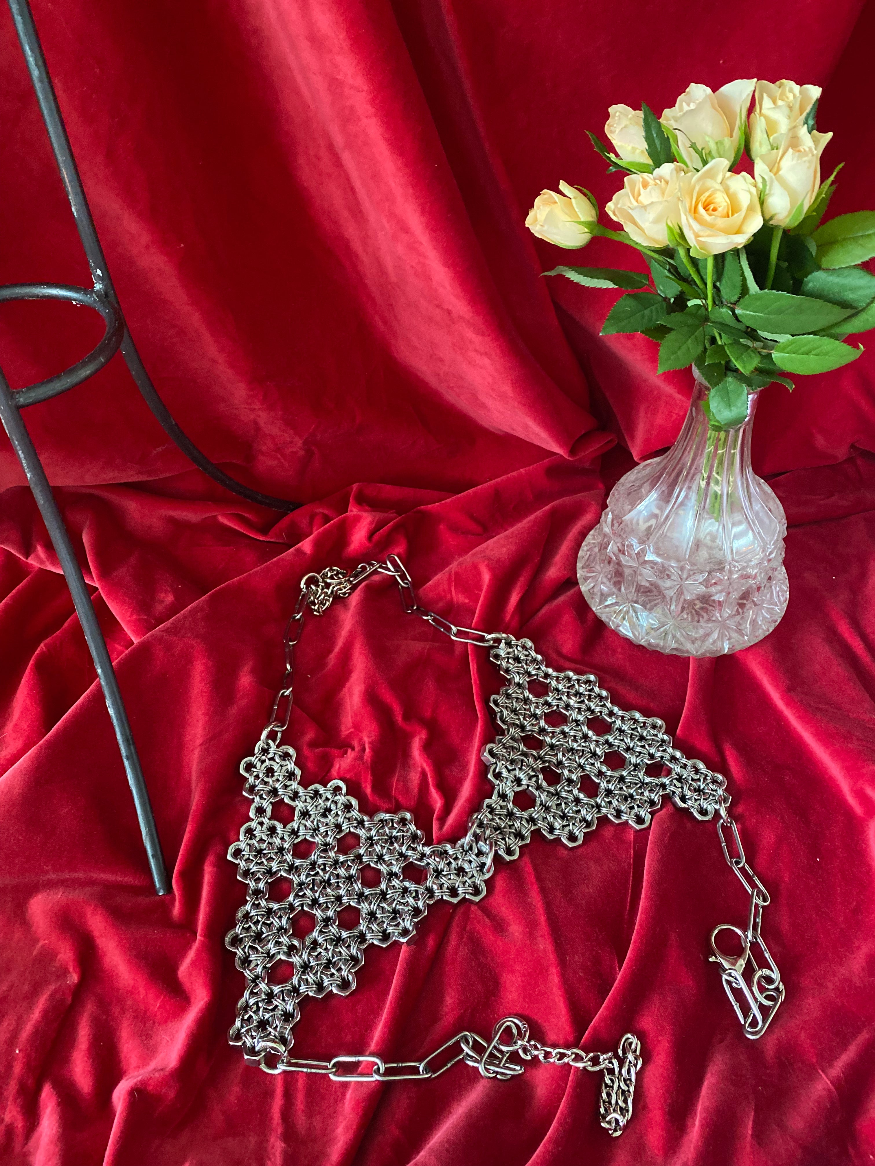 Handmade chainmail bra made from stainless steel rings and industrial screws, displayed on wine-red fabric with a flower, creating a gothic alternative fashion look. Made in Berlin, featuring a unique shiny silver armor-inspired design for statement-making style.
