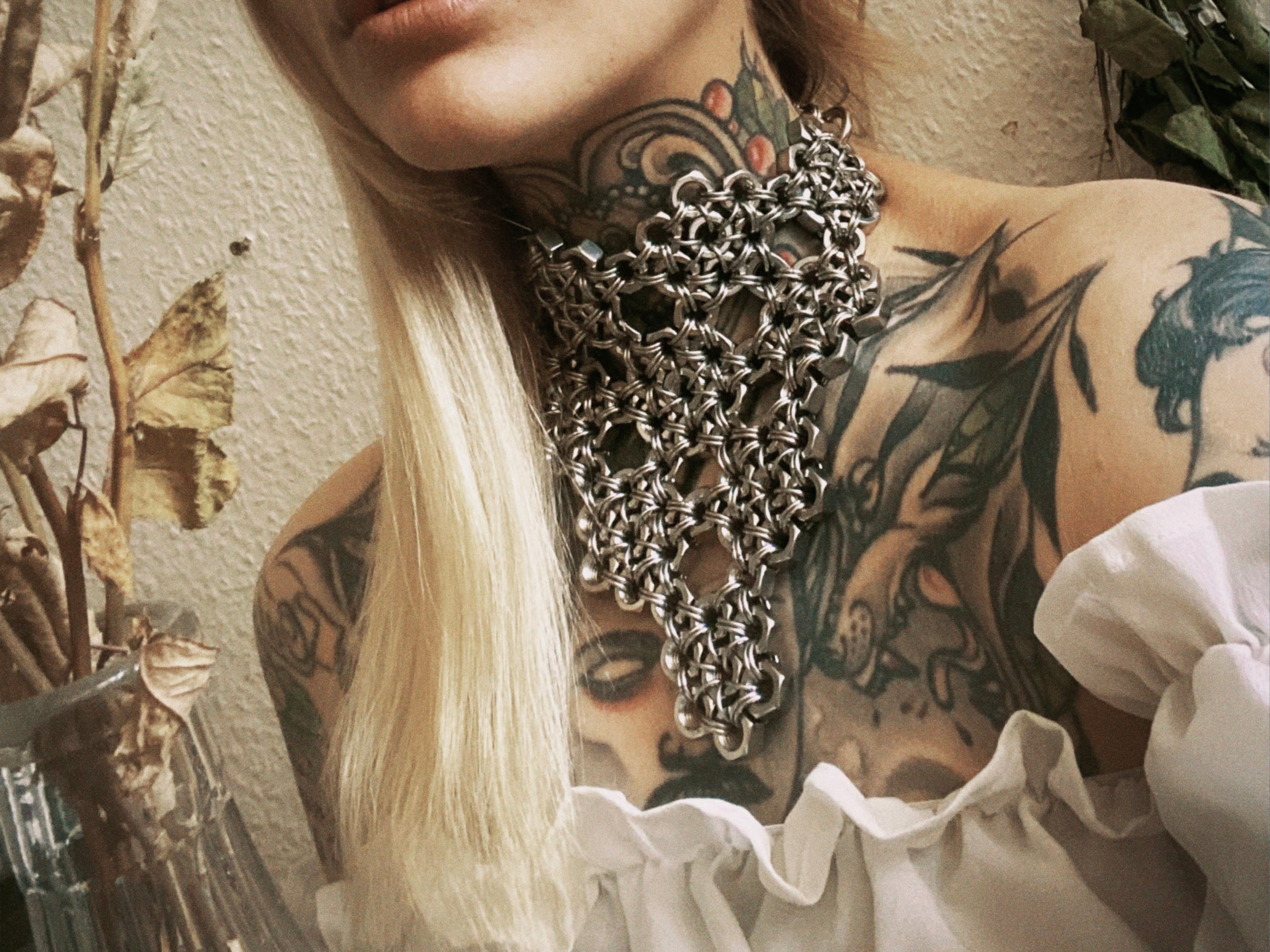 Close-up of a blonde tattooed model with neotraditional tattoos wearing a handcrafted industrial chainmail armor necklace. Made in Berlin from stainless steel rings and screws, featuring a unique, never-before-seen chainmail pattern. Statement piece for alternative fashion.