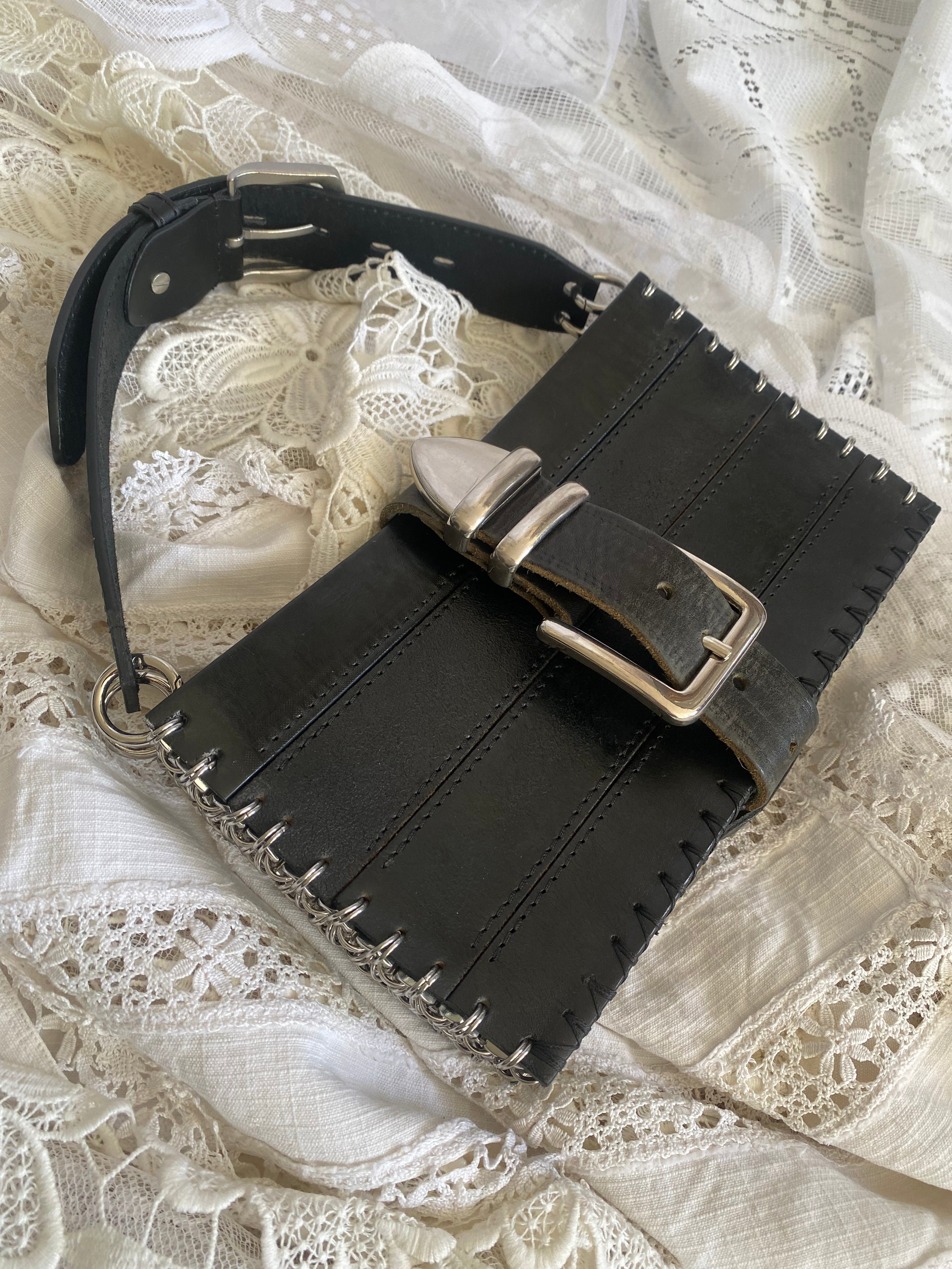 Limited edition handmade black leather handbag from upcycled materials, lying flat with shiny chainmail detail on the side. Can also be worn as a shoulder bag. An avant-garde statement piece photographed against a gothic Victorian aesthetic background.