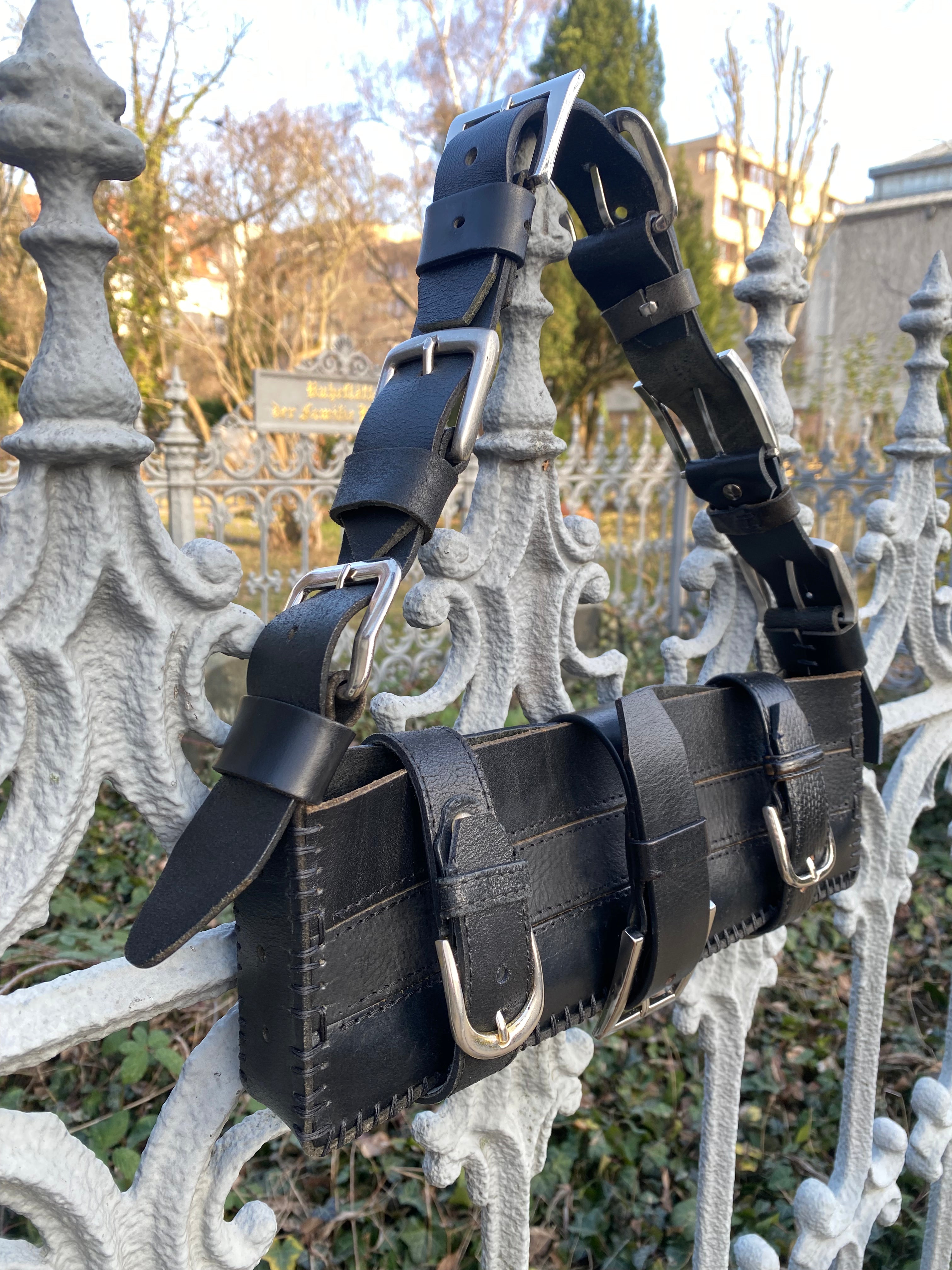Black upcycled and sustainable leather handbag hanging from a white Baroque-style fence, featuring a handle made of interconnecting belt buckles and leather pieces.