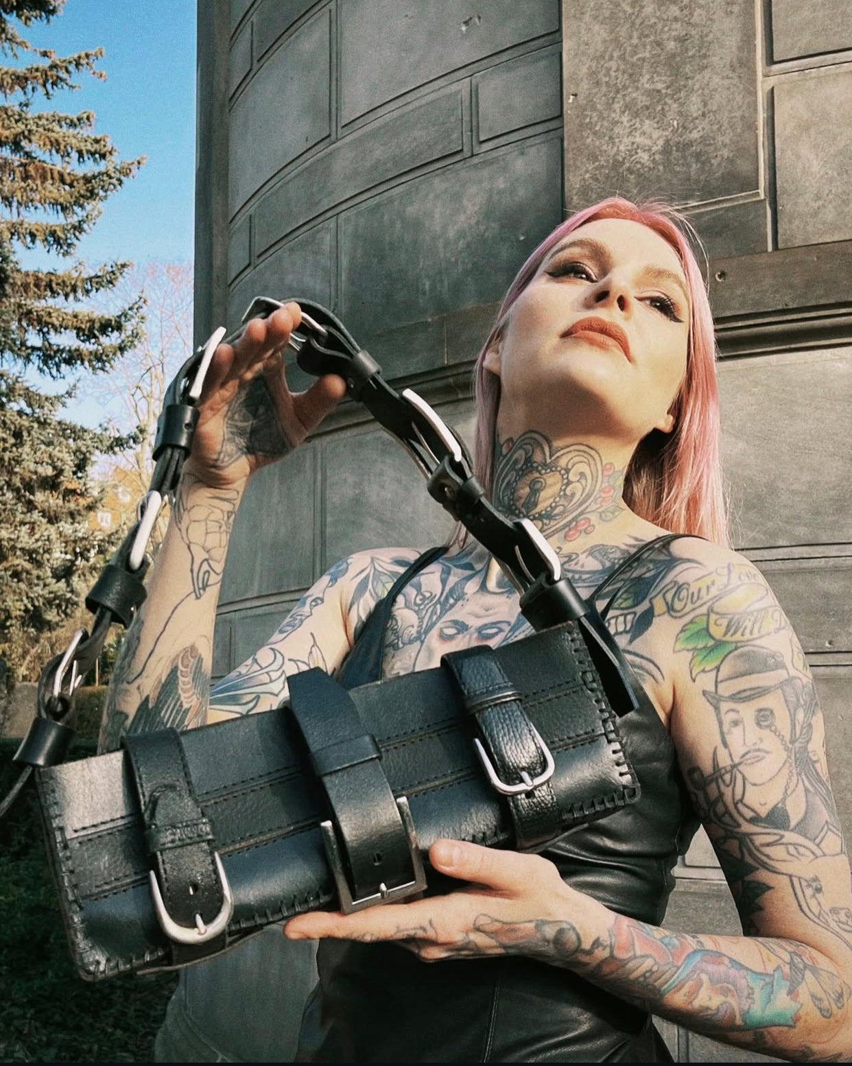 Tattooed pink-haired model holding a unique black leather handbag made from repurposed materials. Long rectangular bag with three buckle closures, designed as a classic black leather handbag that can also be worn as a shoulder bag. Features belt buckle handle and metallic details for a bold, sustainable fashion look.